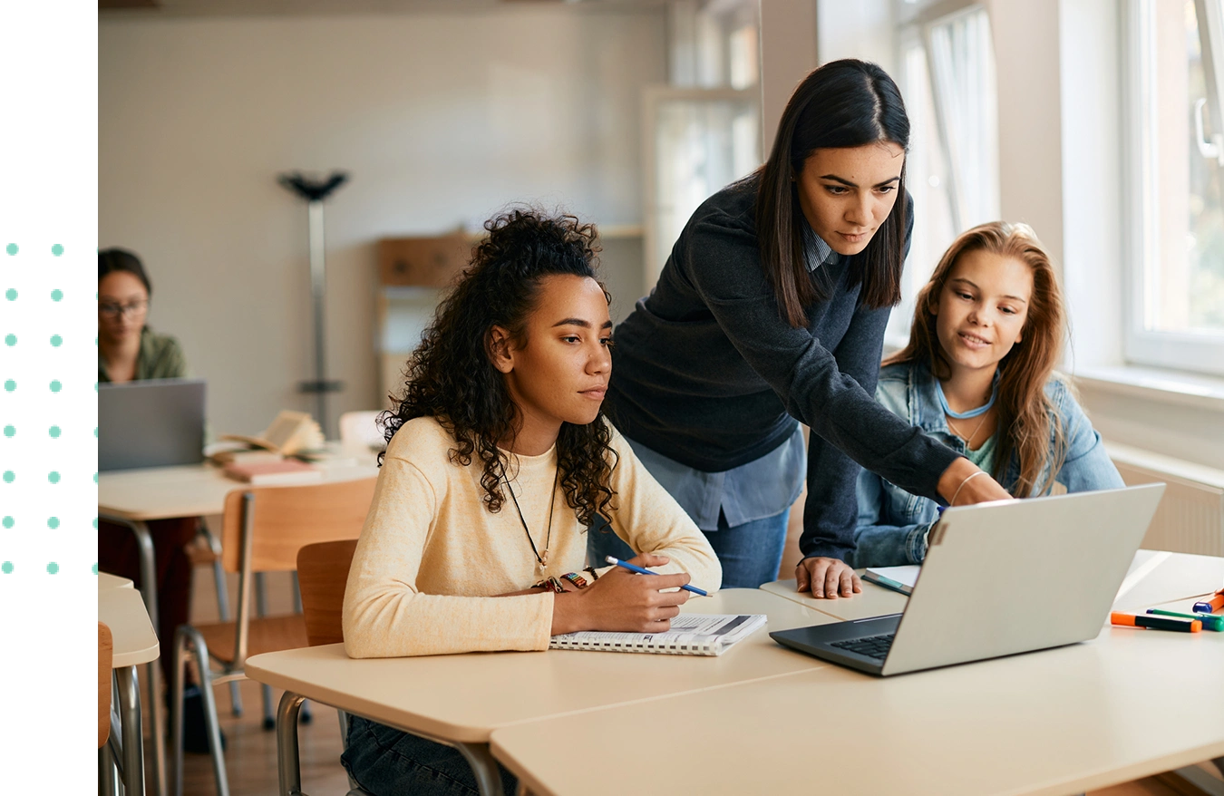 A teacher assists two students working on a laptop at a classroom table.