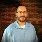 Ray-M.-Sanchez A man in glasses and blue shirt standing next to brick wall.